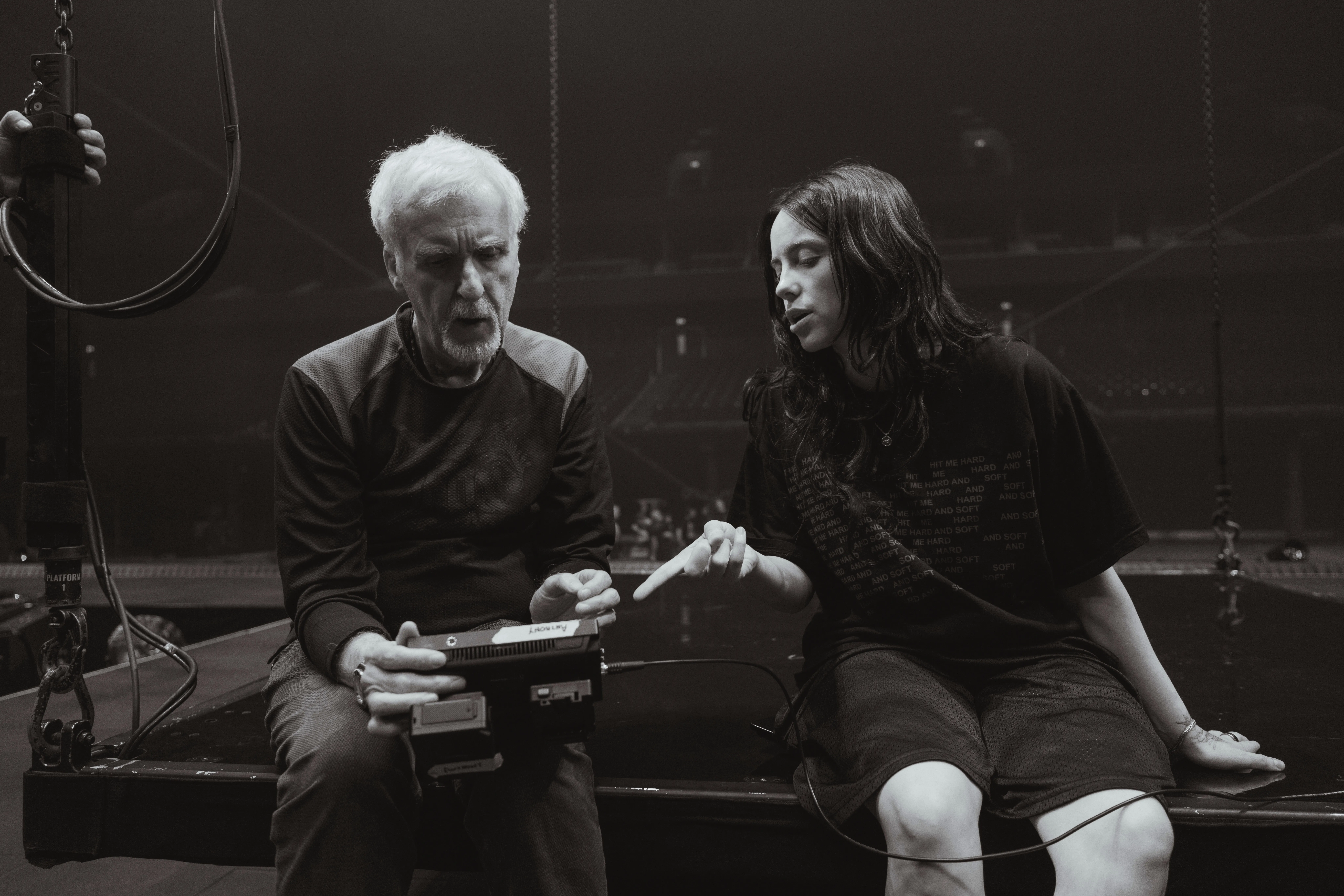 James Cameron and Billie Eilish sitting together on the edge of the stage, reviewing footage on a small handheld 3D monitor.