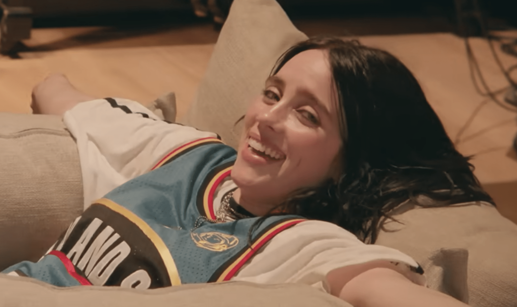 Billie Eilish laughing while reclined on a couch in a vintage Memphis Grizzlies jersey, from one of the film's documentary interview segments.