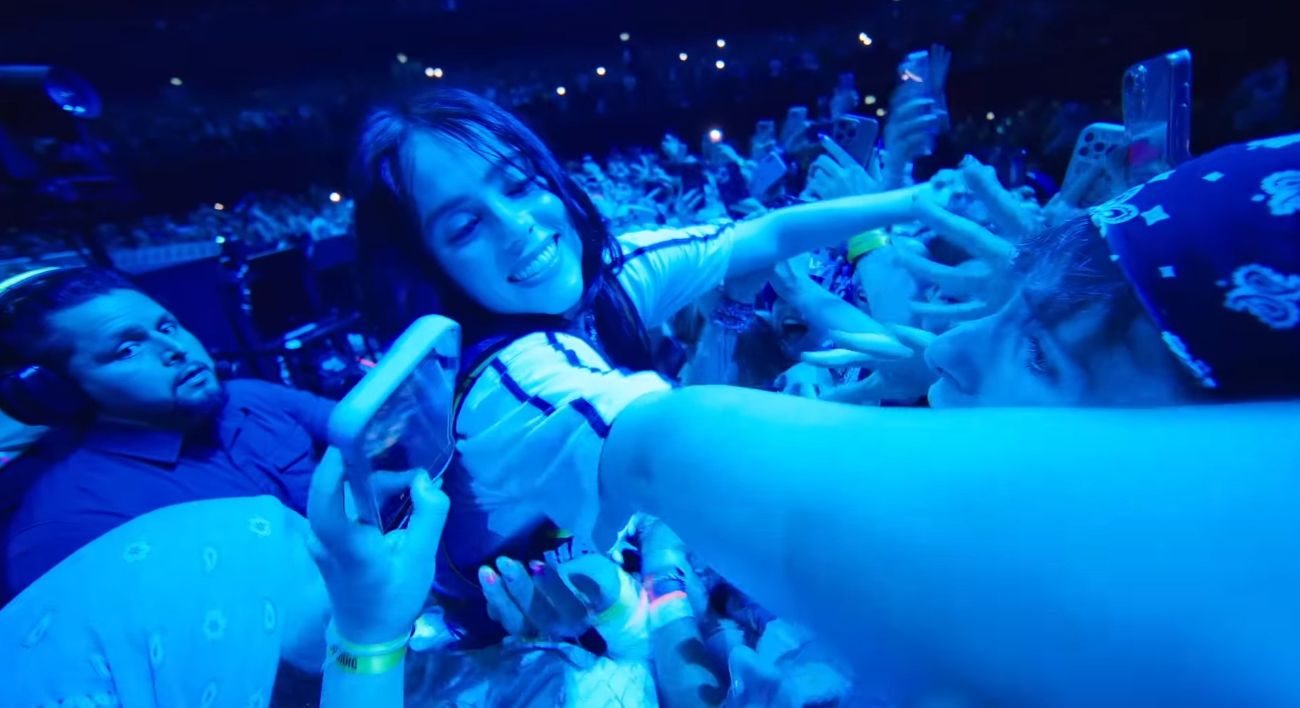 Billie Eilish in the pit surrounded by fans holding up phones during the high-five run, lit in deep blue.