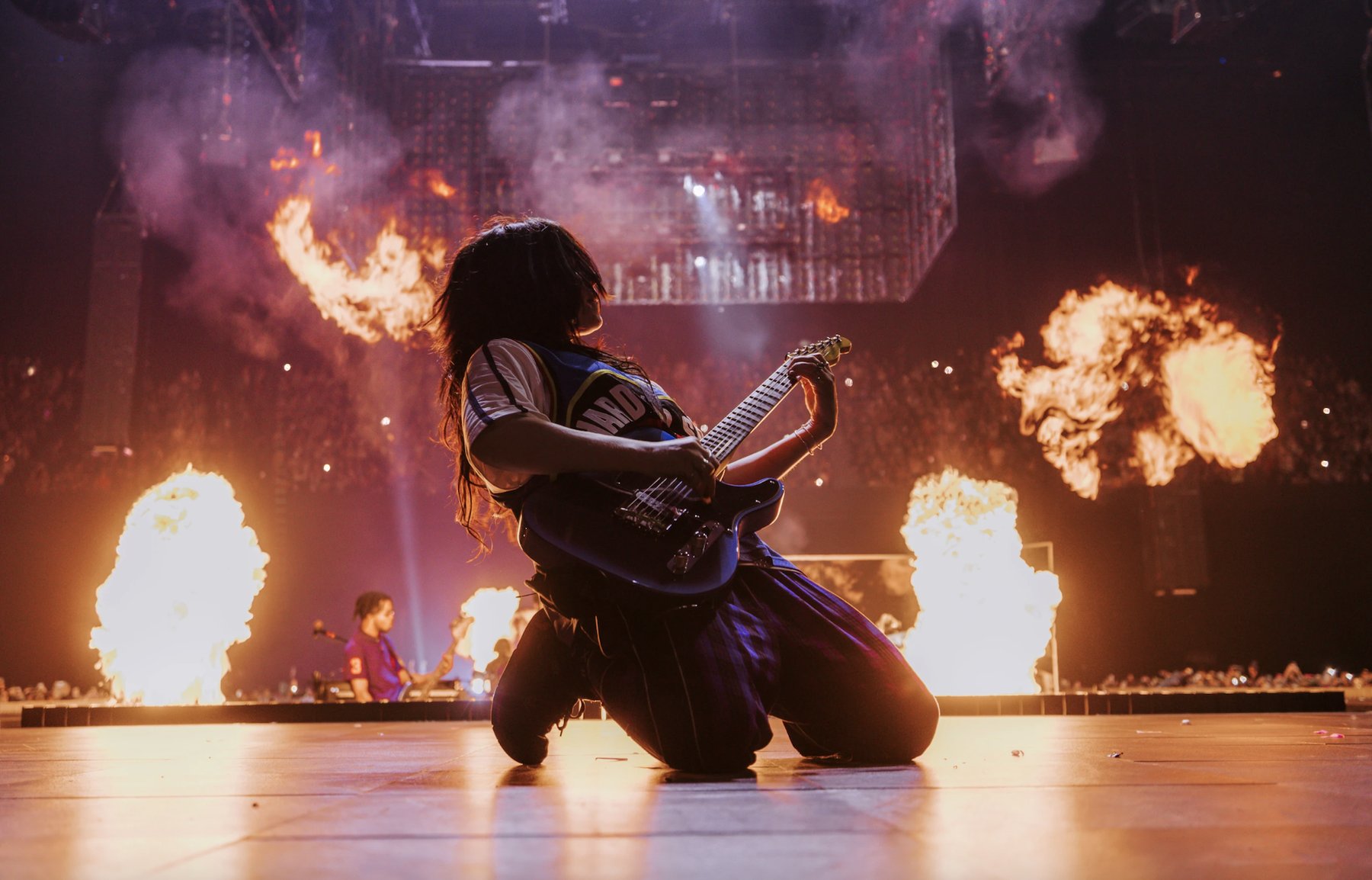 Billie Eilish kneeling on stage playing electric guitar, flanked by columns of stage fire, during Hit Me Hard and Soft: The Tour.